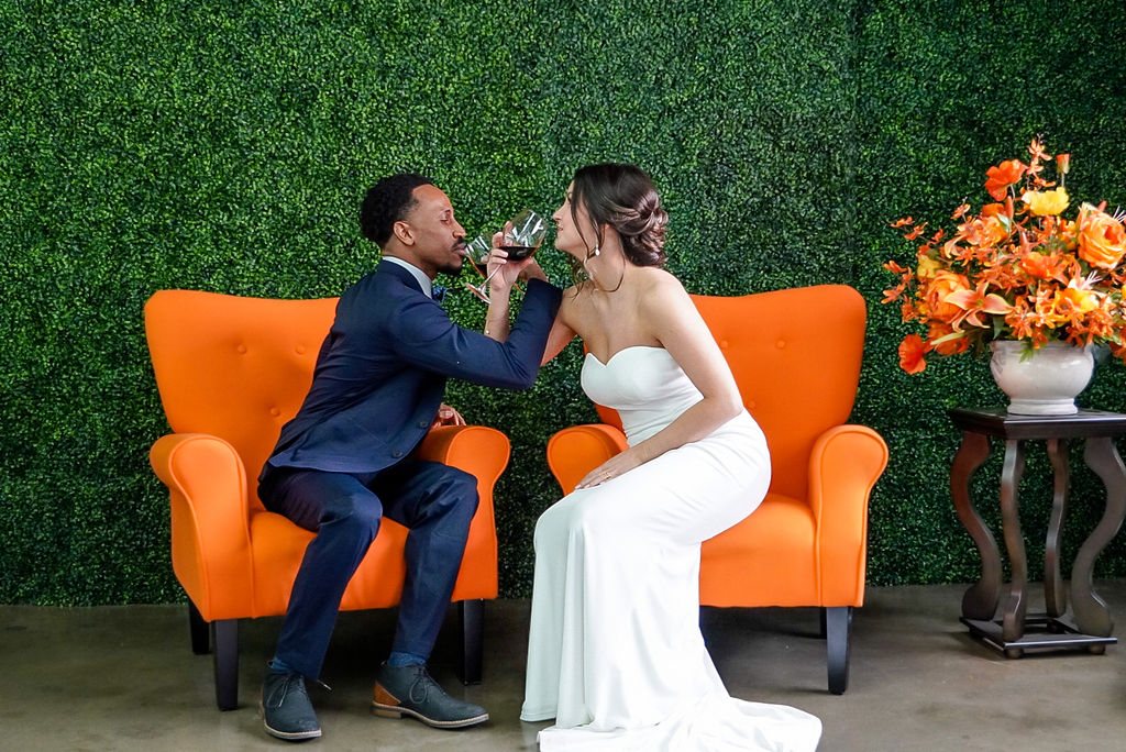 A couple toasting in orange armchairs against the greenery wall.