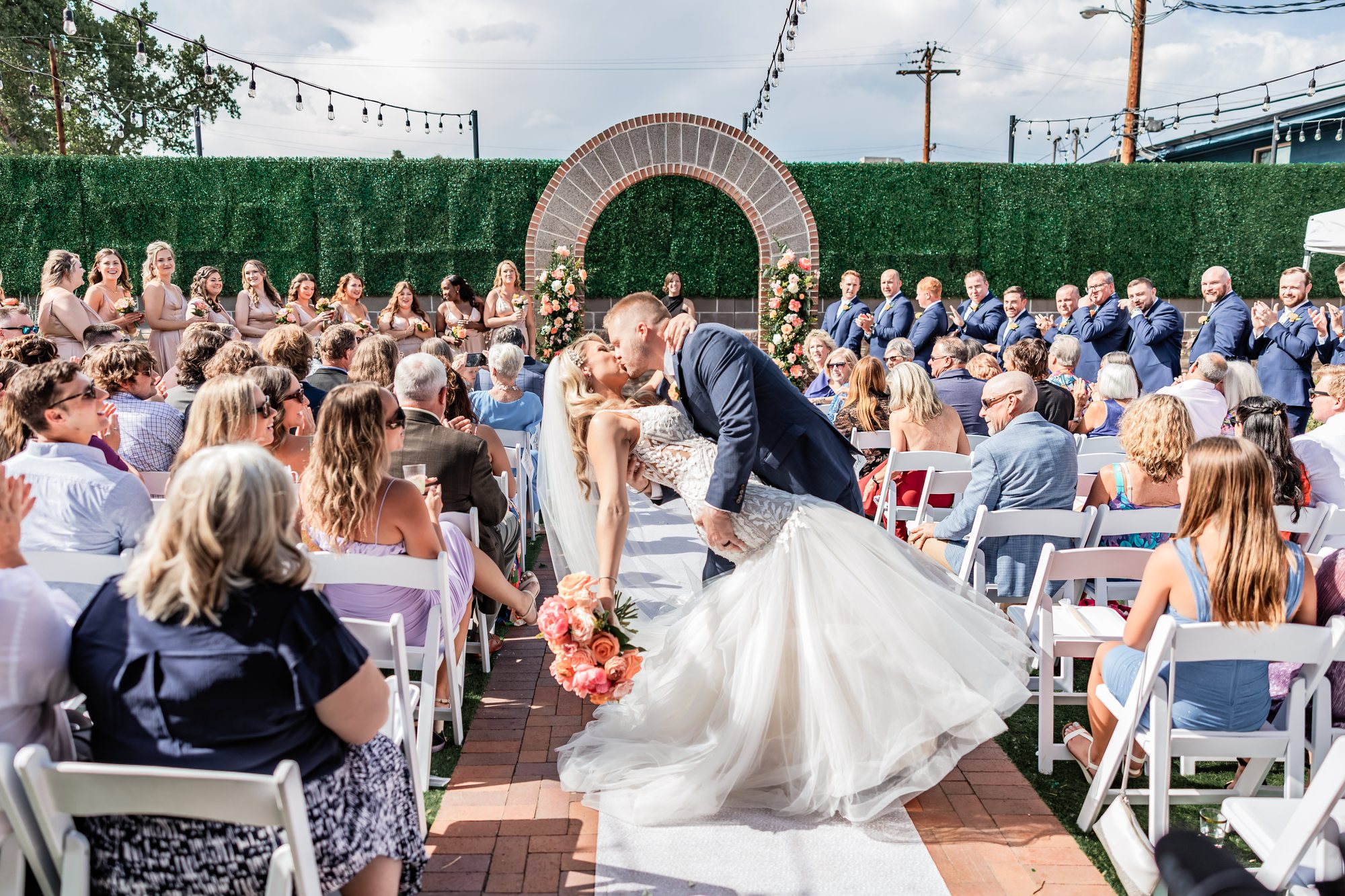 An outdoor ceremony under the brick arch, the couple kissing as guests applaud.